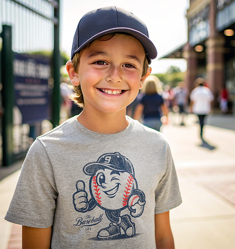 A young boy at a baseball stadium, wearing a heather gray t-shirt with a cartoon baseball mascot and a navy baseball cap.