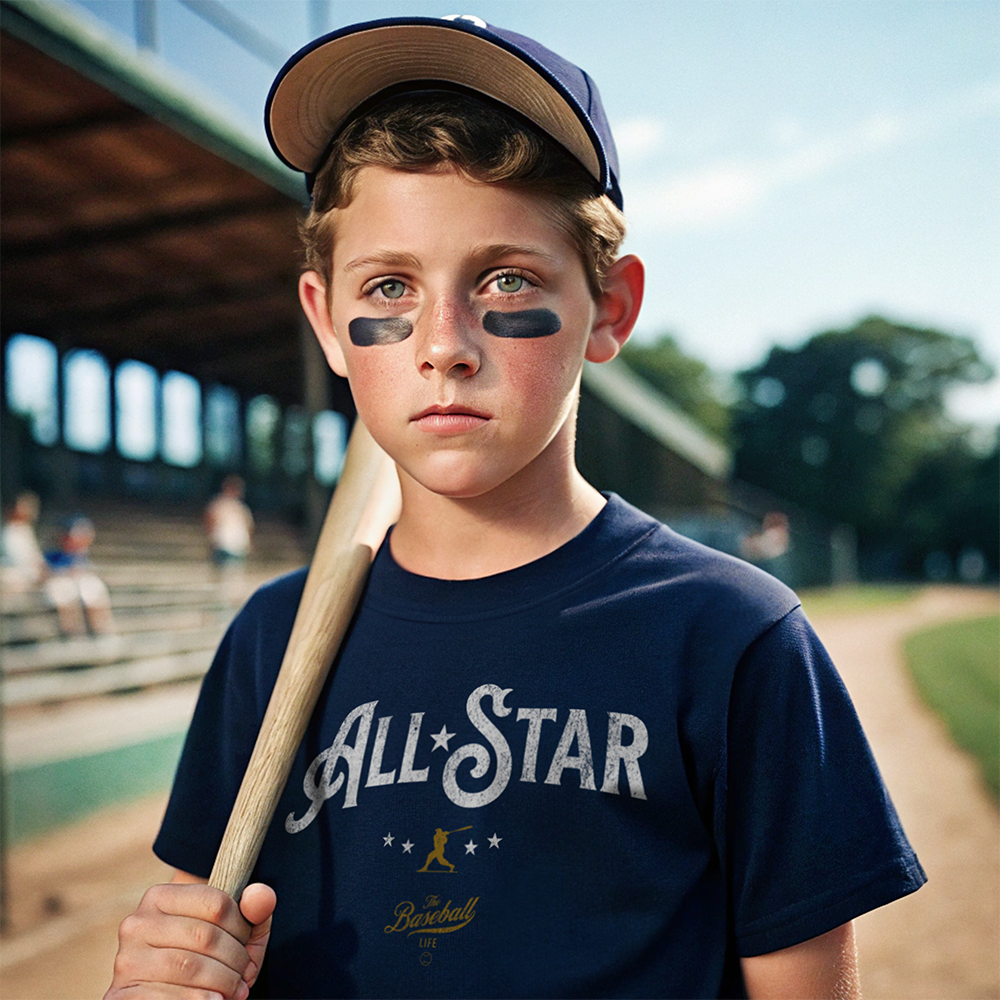 A youth baseball player wearing a navy All-Star t-shirt, with a baseball bat over his shoulder, standing on a baseball field.