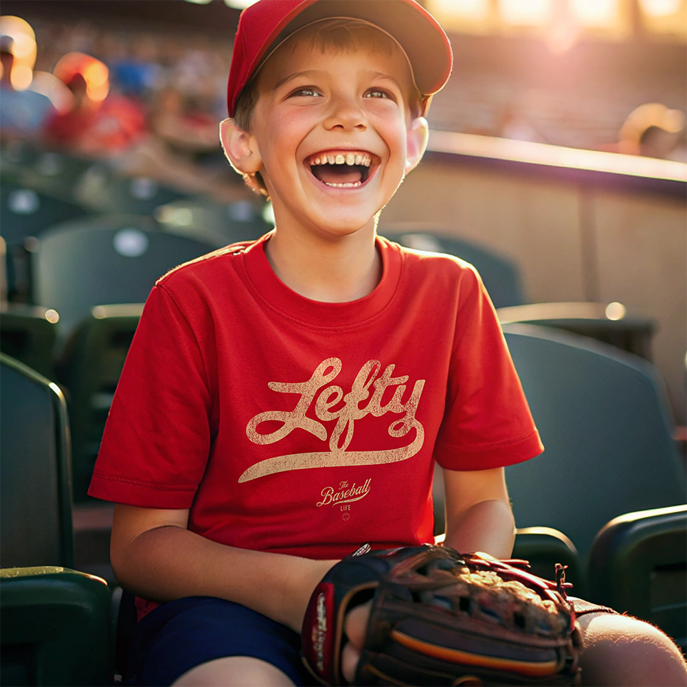 A young boy at a baseball game, wearing a red t-shirt with the word "Lefty" across the chest.