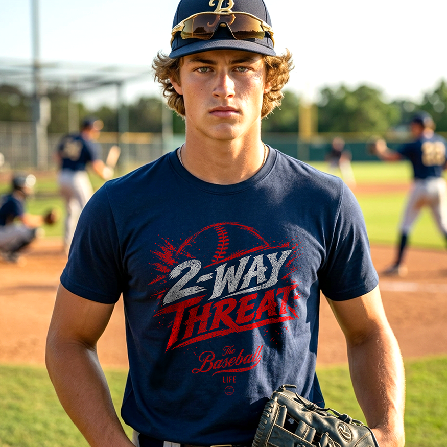 A baseball player wearing a navy baseball t-shirt featuring a high-energy "2-WAY THREAT" graphic with The Baseball Life logo, standing on baseball field.